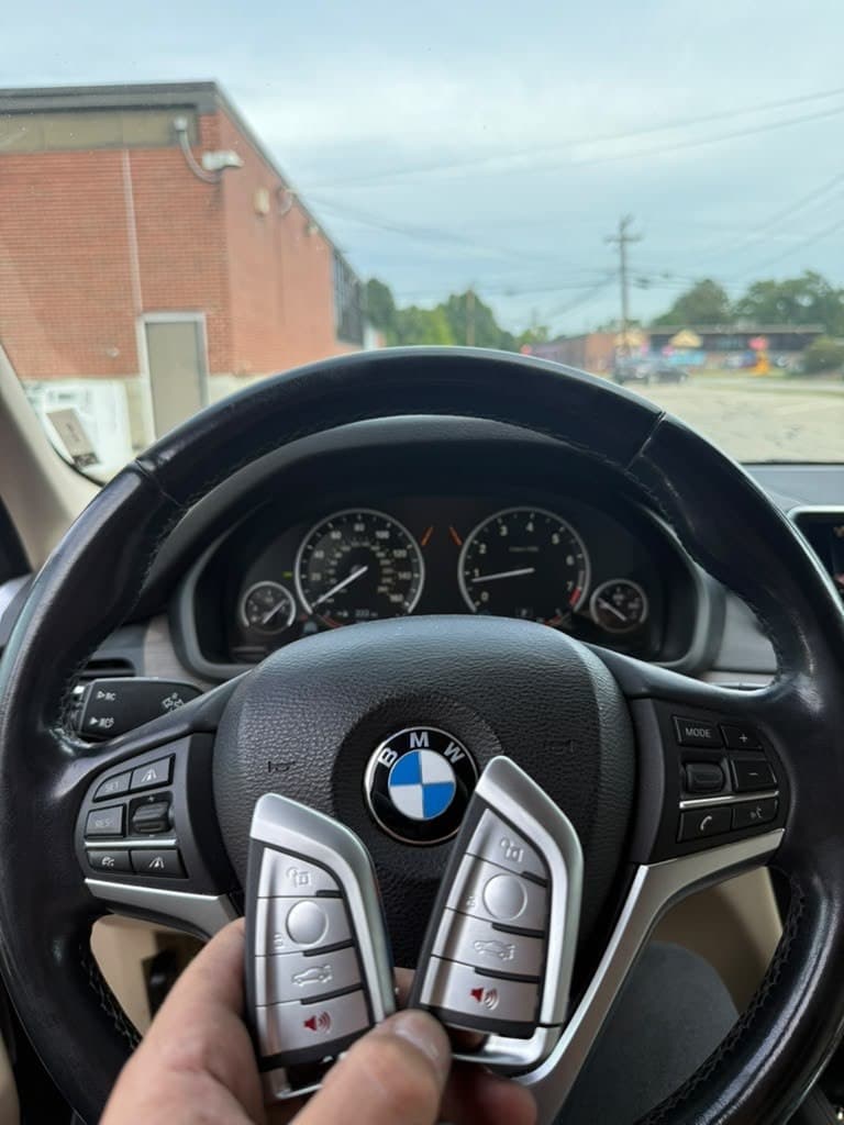 Hand holding two silver BMW key fobs in front of a BMW steering wheel.