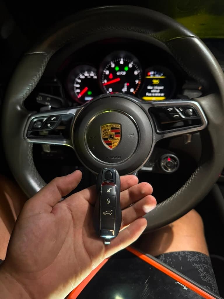 Hand holding a key fob in front of a Porsche steering wheel and illuminated dashboard.