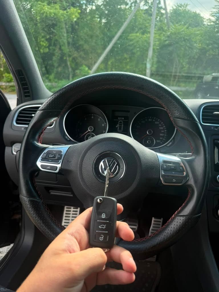 Hand holding a Volkswagen key fob in front of a steering wheel with red stitching.