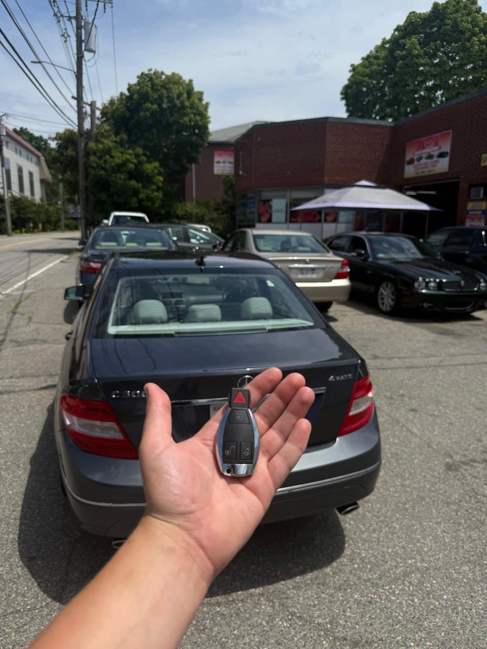 Hand holding a Mercedes-Benz key fob in front of a grey C300 sedan outdoors.